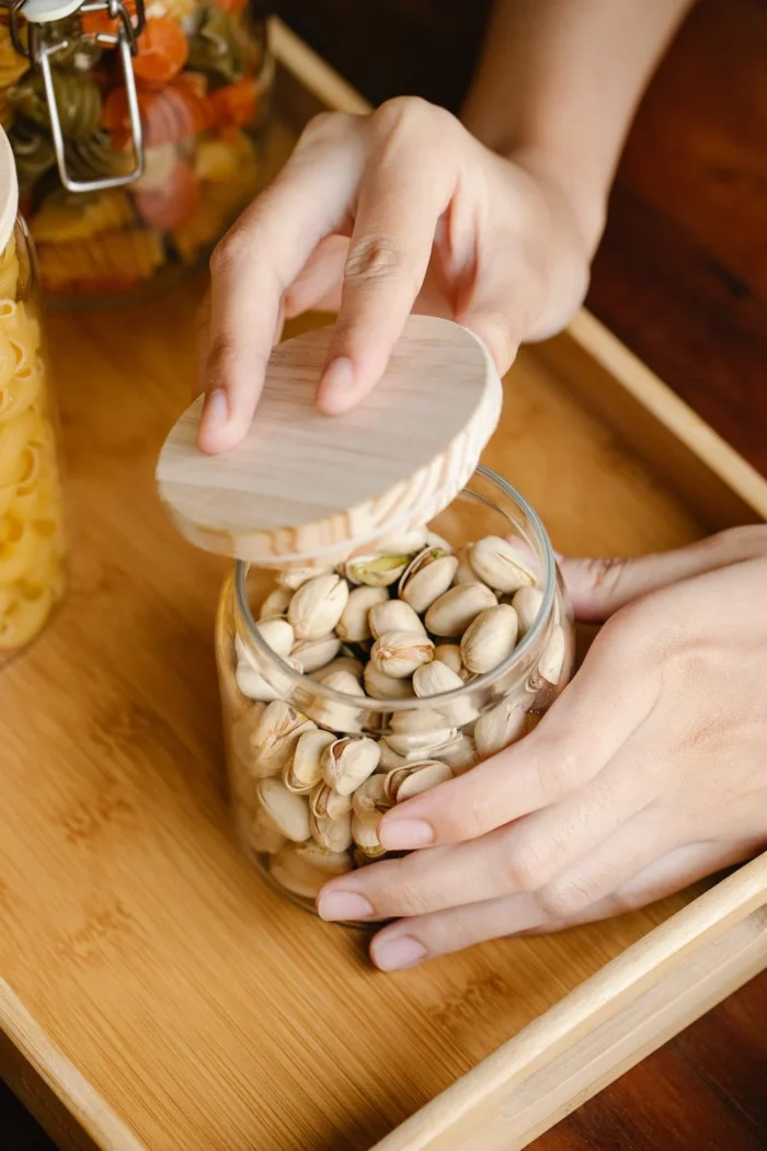 A jar full of pistachios standing on a wooden tray, with hands shown closing a bamboo lid