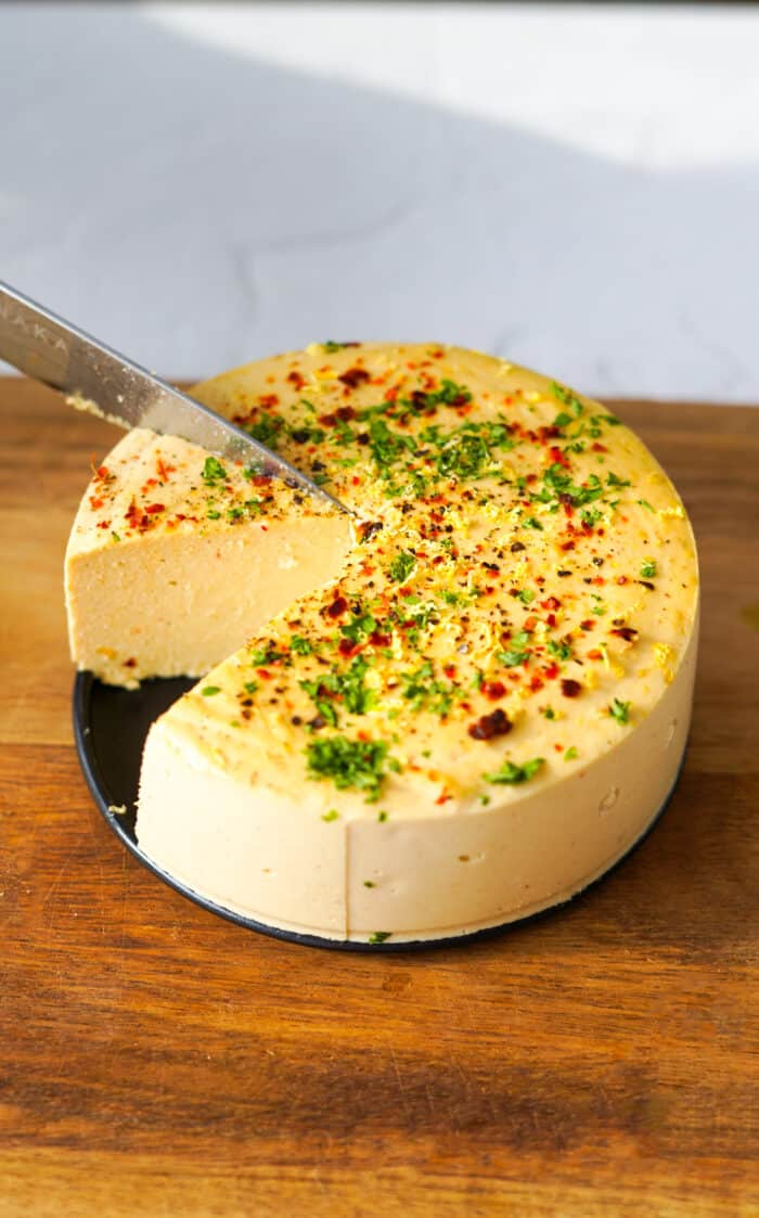 a knife slicing into a round block of vegan hard cheese on a cutting board