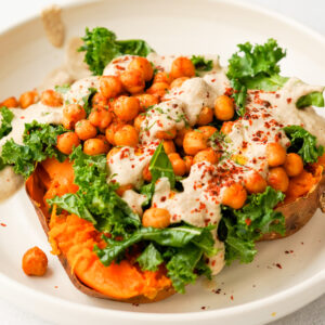 baked sweet potato topped with roasted spiced chickpeas, kale, and creamy sunflower sauce, served in a white bowl