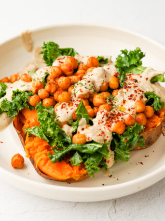 baked sweet potato topped with roasted spiced chickpeas, kale, and creamy sunflower sauce, served in a white bowl
