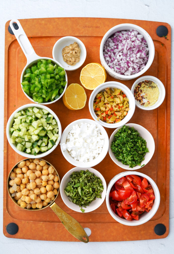 ingredients for the vegan greek pasta salad arranged in individual bowls on top of a wooden cutting board