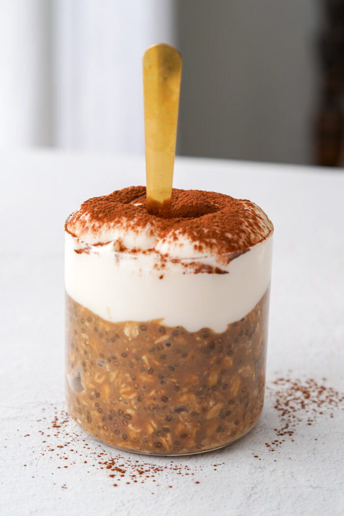 a clear jar of overnight oats with yogurt and cocoa powder over top and a gold spoon sticking out