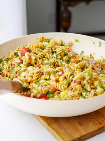 vegan greek pasta salad mixed together in a large white mixing bowl on top of a wooden cutting board