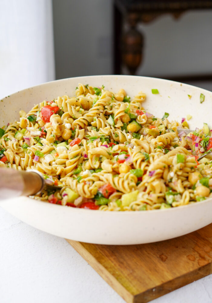 vegan greek pasta salad mixed together in a large white mixing bowl on top of a wooden cutting board