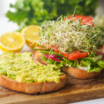 an open faced sandwich open on a cutting board with smashed avocado on one side and lettuce, tomato and sprouts on the other