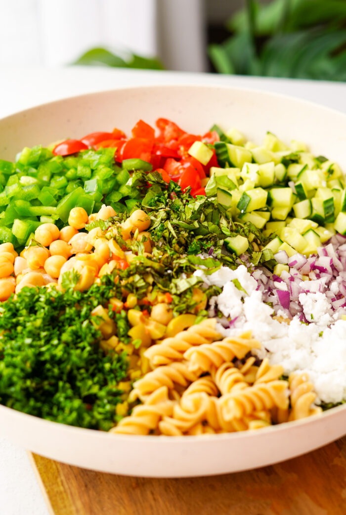 ingredients of the greek pasta salad arranged in a large white mixing bowl