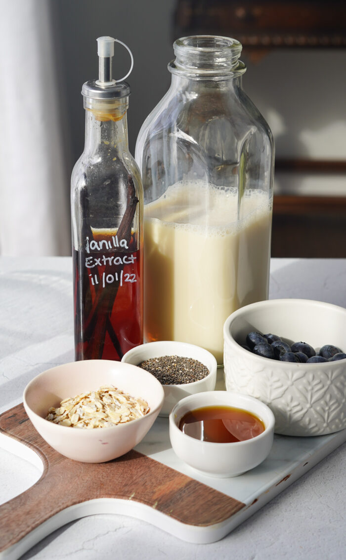 a whole bunch of ingredients including vanilla extract, maple syrup, plant-based milk, oats and blueberries sitting on a counter top