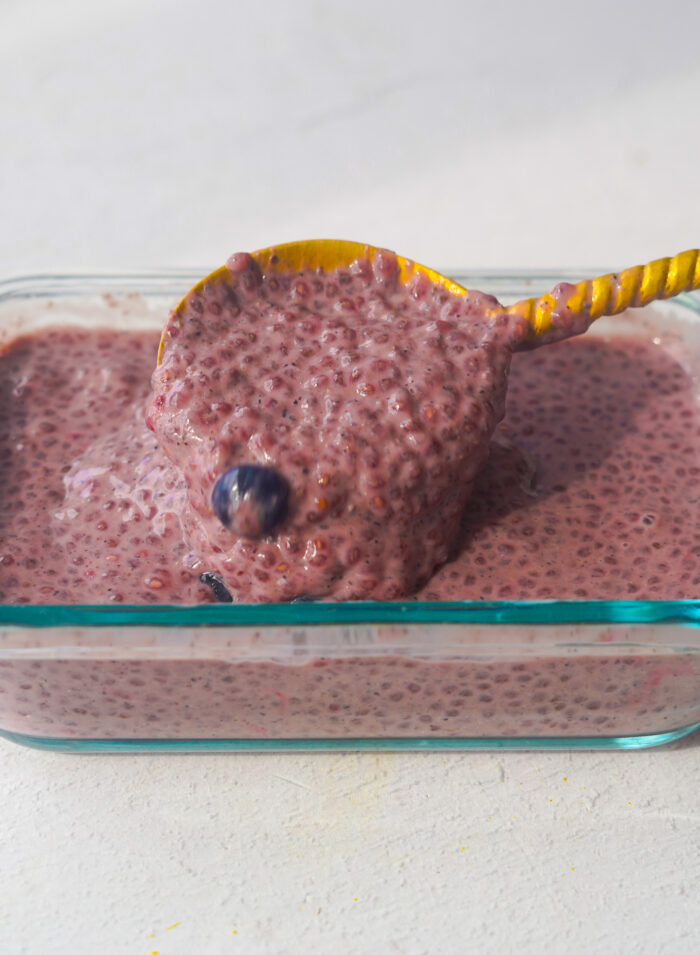 blueberry chia pudding in a Tupperware glass container with a gold spoon dripping off