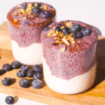 two blueberry chia pudding cups on a wood cutting board with granola and blueberries on top