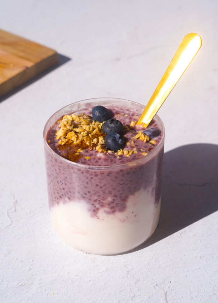 a blueberry chia pudding parfait with a gold spoon sticking out sitting on a white backdrop