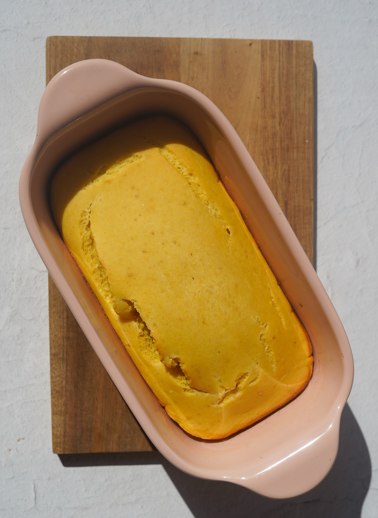 vegan lemon loaf freshly out of the oven, in a pastel ceramic loaf pan on top of a wooden cutting board