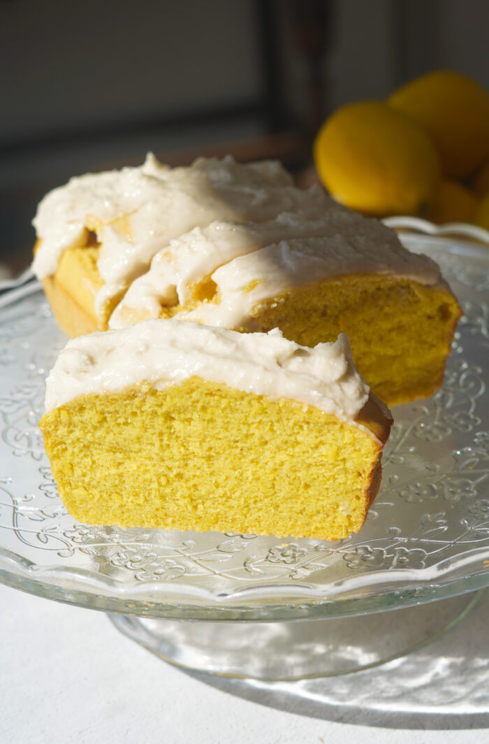 vegan lemon loaf on a glass cake stand, sliced and iced with vanilla frosting