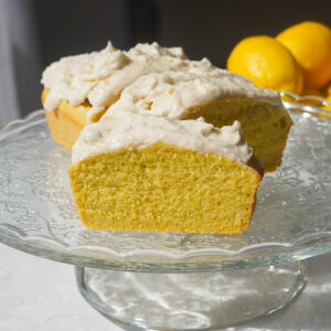 A glass cake stand holding a yellow lemon loaf with vanilla frosting, with lemons in the background