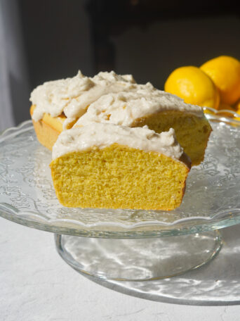 A glass cake stand holding a yellow lemon loaf with vanilla frosting, with lemons in the background