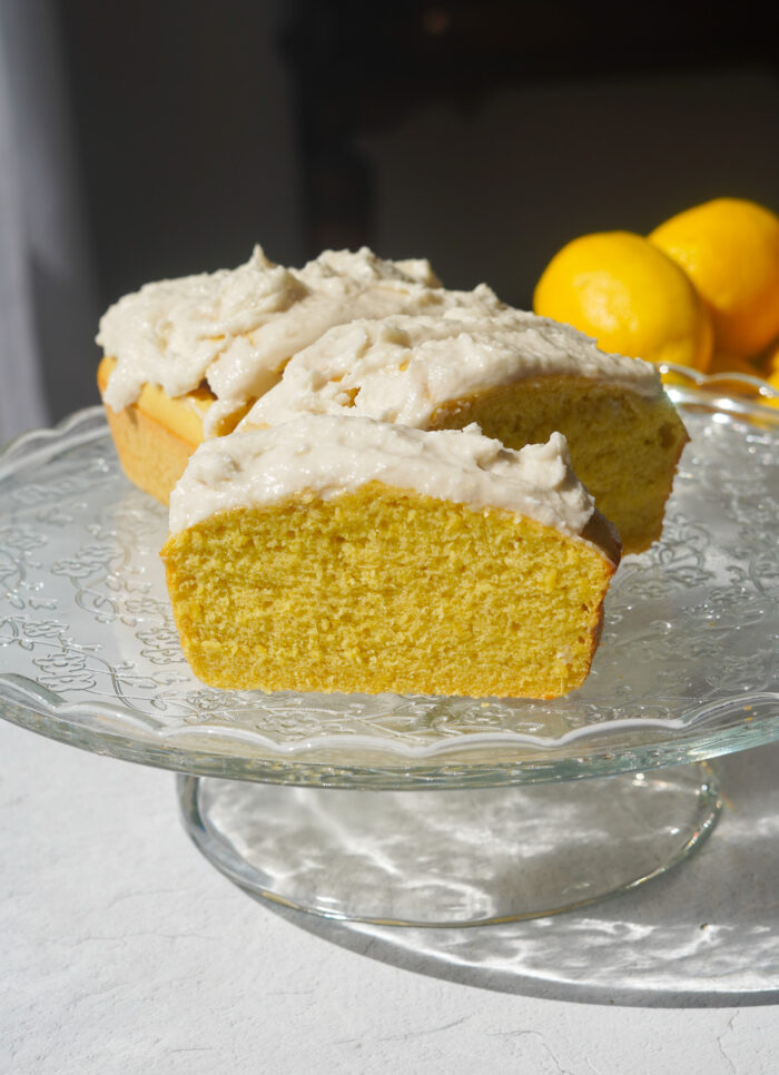 A glass cake stand holding a yellow lemon loaf with vanilla frosting, with lemons in the background