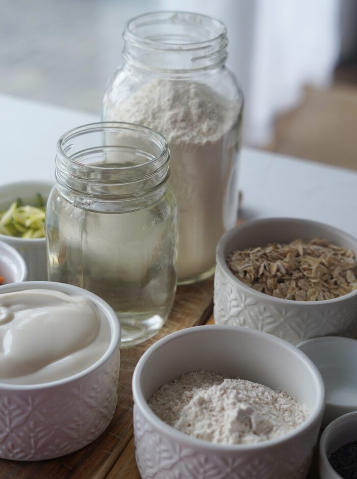 ingredients for overnight oat buns set out on a wooden board, including vegan yogurt, water, flour, zucchini, oats, and carrots