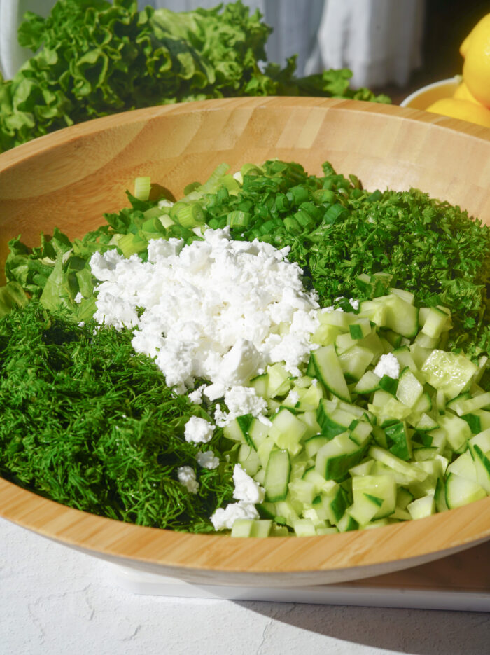 cucumber, parsley, dill, vegan feta, green onions and lettuce sitting in a wooden bowl on a white counter