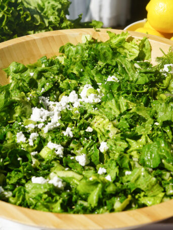 cucumber, parsley, dill, vegan feta, green onions and lettuce sitting in a wooden bowl on a white counter