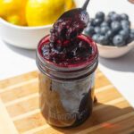 blueberry jam sitting in a small jar on a wood cutting board with a spoon in it