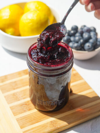 blueberry jam sitting in a small jar on a wood cutting board with a spoon in it