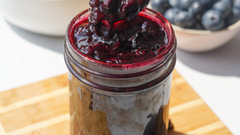 blueberry jam sitting in a small jar on a wood cutting board with a spoon in it