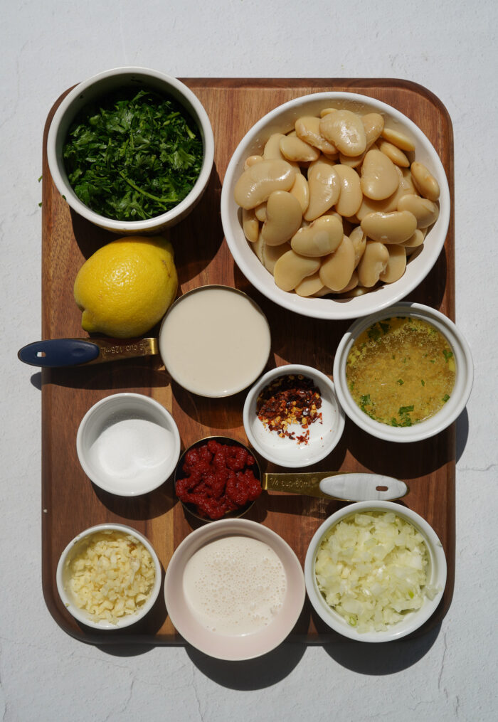 a brown cutting board on a white backdrop with creamy butter bean ingredients including a bowl of beans, parsley, tomato paste and nutritional yeast