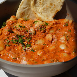 a grey pan full of butter beans in a red sauce with a naan at the back