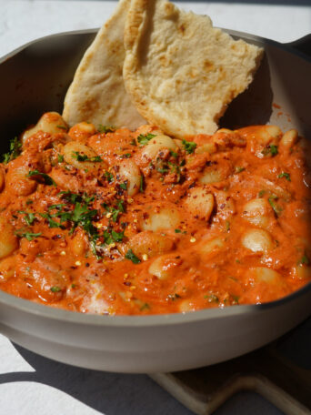a grey pan full of butter beans in a red sauce with a naan at the back