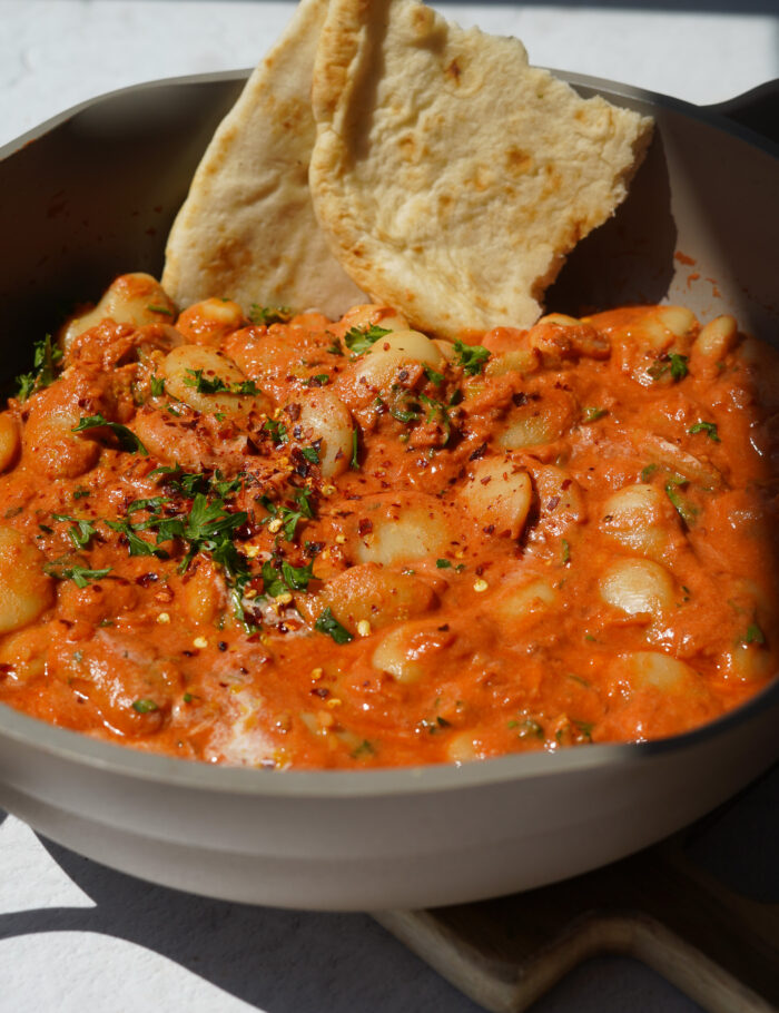 a grey pan full of butter beans in a red sauce with a naan at the back 