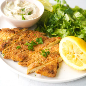 vegan schnitzel on a white plate with a squeezed lemon, vegan tzatziki and a green salad