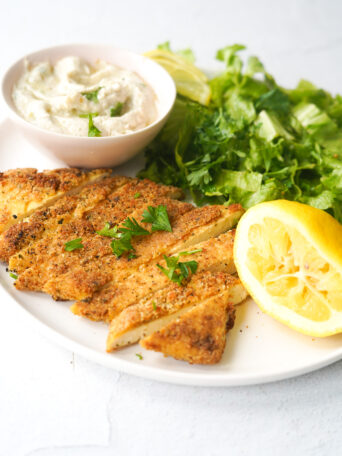 vegan schnitzel on a white plate with a squeezed lemon, vegan tzatziki and a green salad