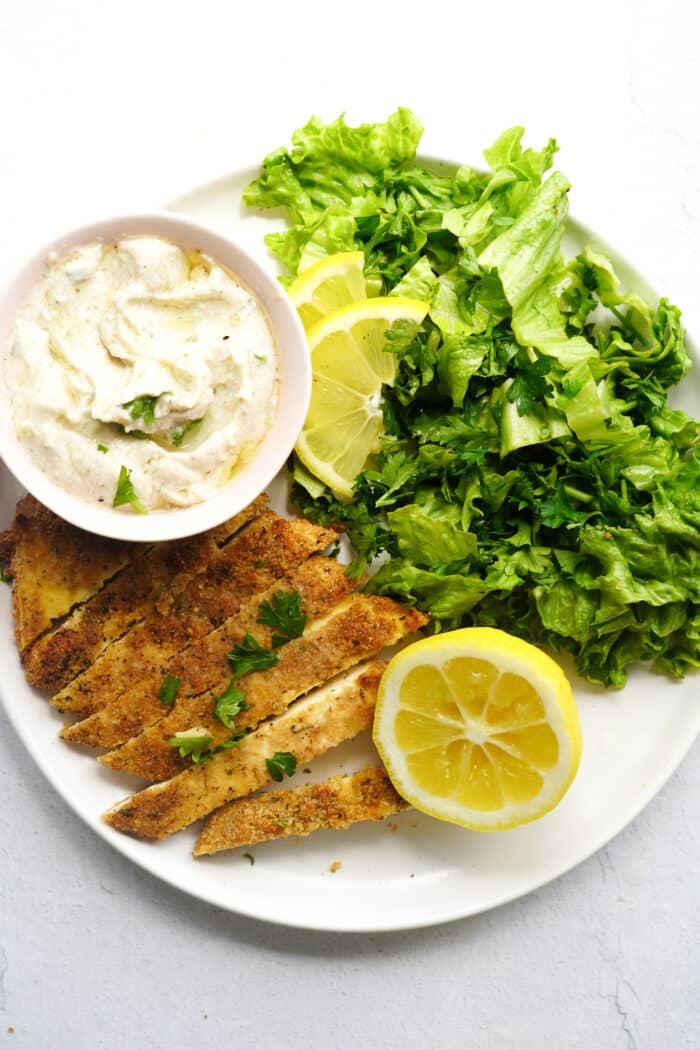 vegan schnitzel on a white plate with a squeezed lemon, vegan tzatziki and a green salad