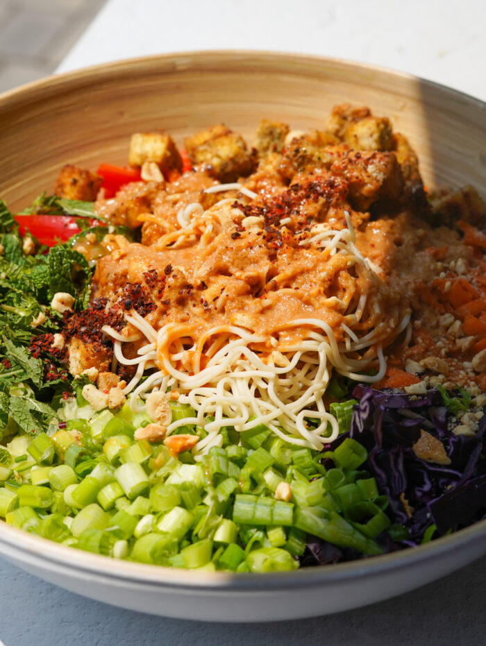 brown bamboo bowl full of tofu, green onion, red cabbage, carrot, noodles, and peanut sauce over top