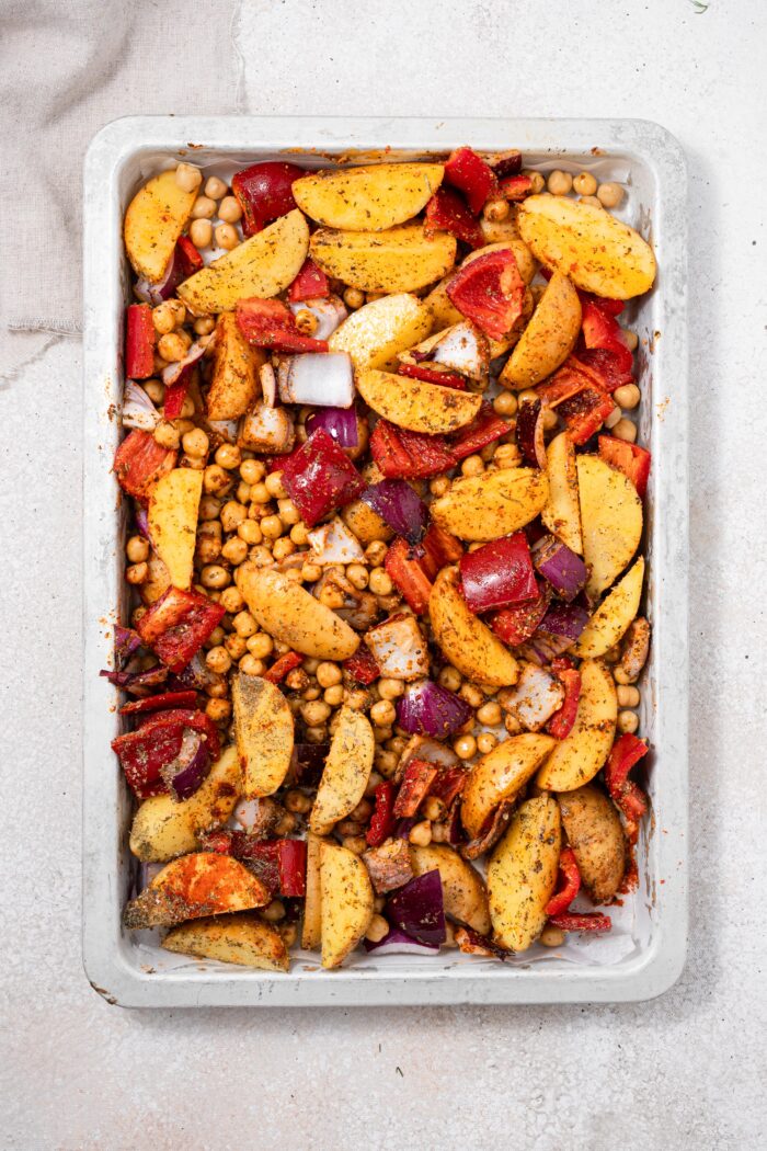 Potatoes, chickpeas, red bell peppers, and red onions on a baking sheet with herbs and spices.