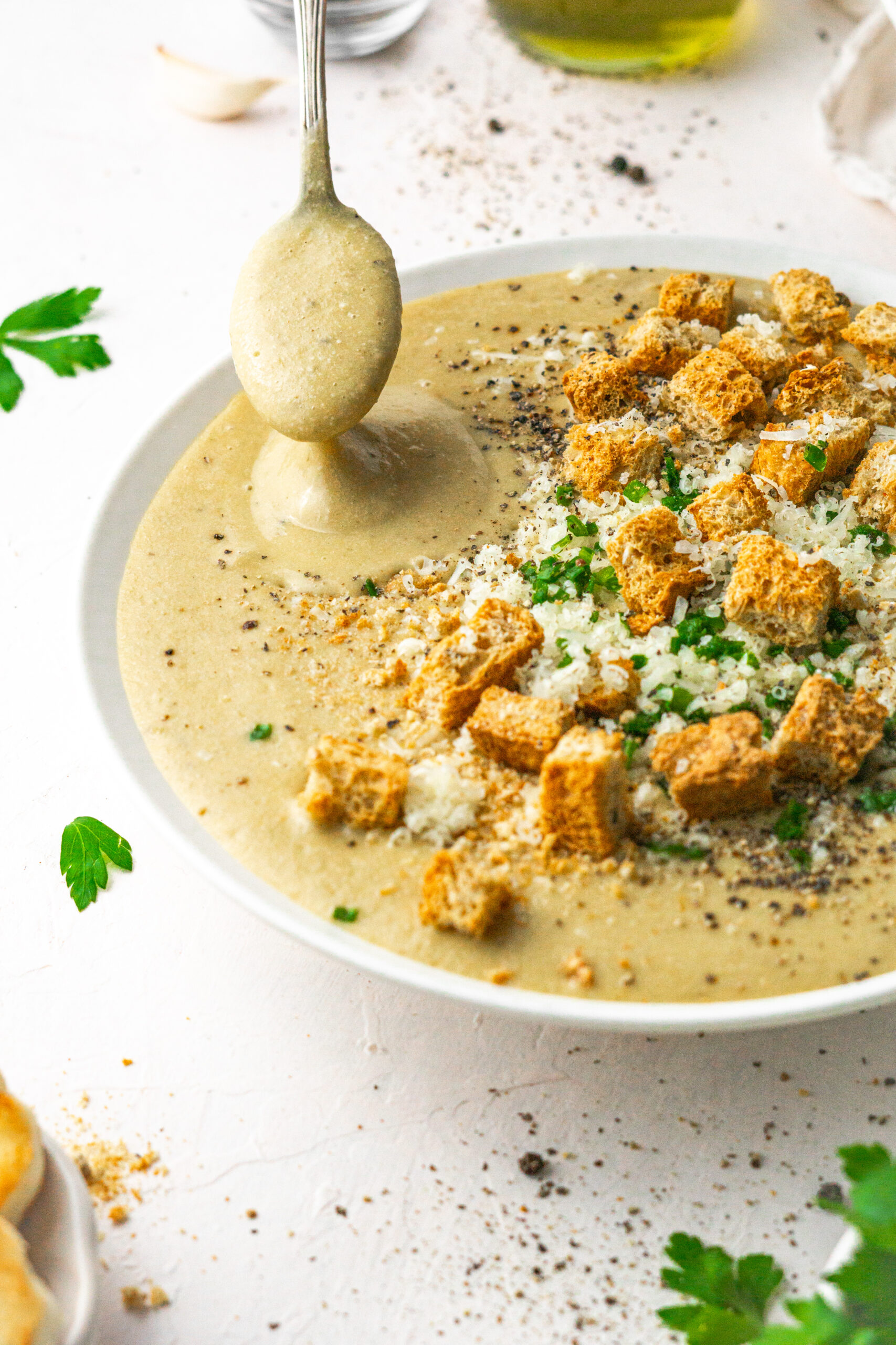 30 clove garlic soup served with choves, vegan parmesan and croutons in a white bowl with a spoon picking up some soup