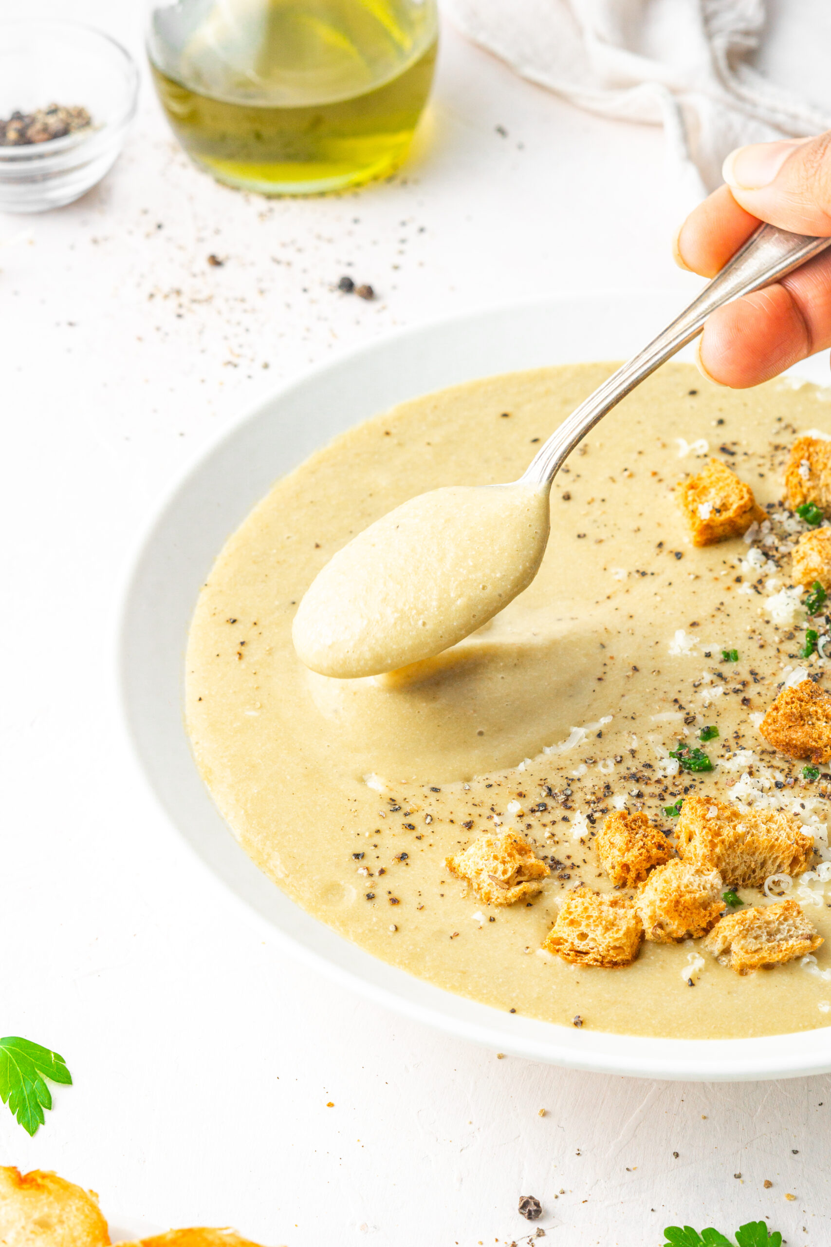 30 clove garlic soup served with choves, vegan parmesan and croutons in a white bowl with a spoon picking up some soup