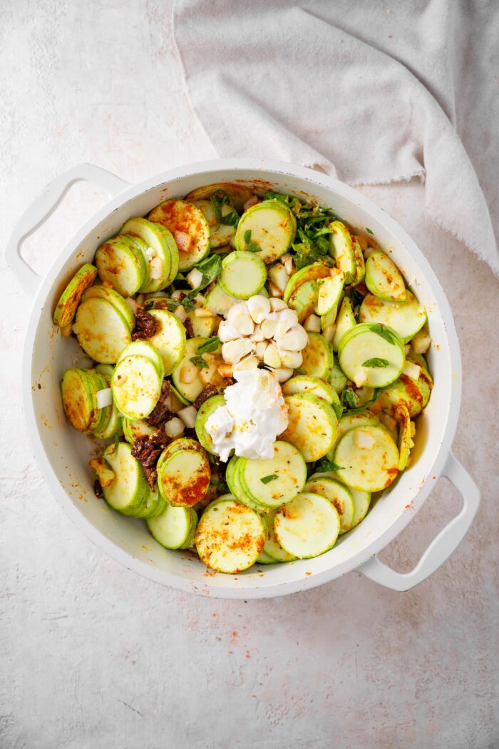 A mixture of vegetables in a white casserole pot, with a whole head of garlic and some vegan cream in the middle.