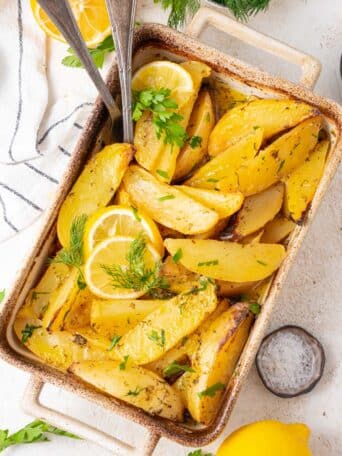 A casserole dish with roasted Greek potatoes with fresh herbs and lemon slices.