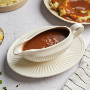 A wide shot of a gravy boat filled with vegan gravy, and mashed potatoes in the background.