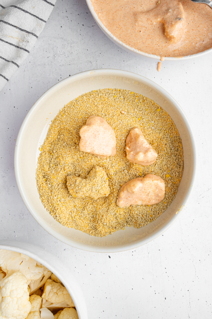 Cauliflower florets dipped in batter and transferred to a bowl with breadcrumbs.