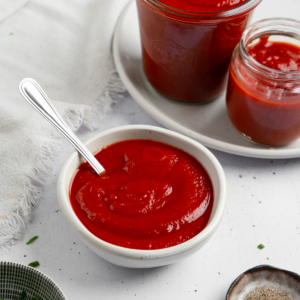 A composed shot of homemade ketchup in a small white bowl with a spoon sticking out, more ketchup in the background.