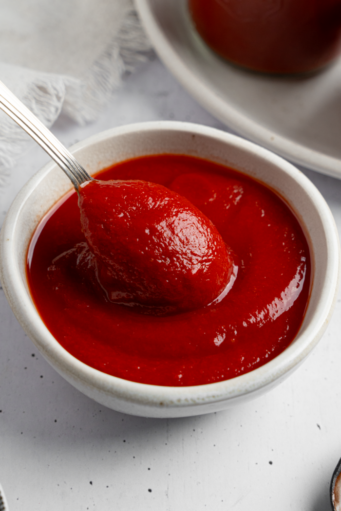 A spoon lifting some homemade ketchup out of a small white ceramic bowl.