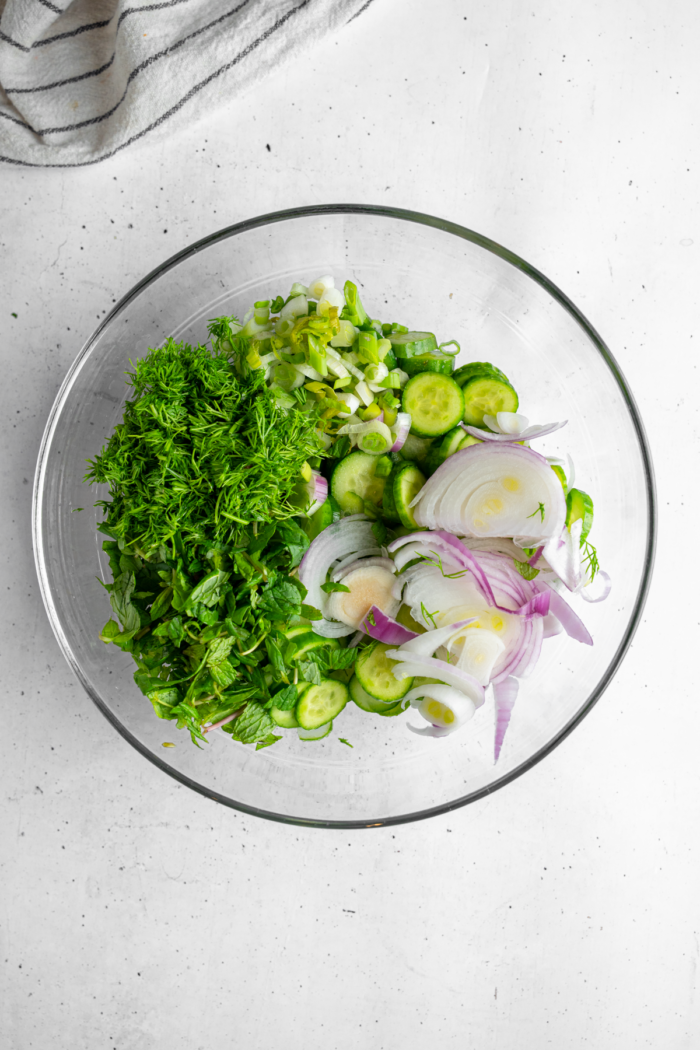 The vegetable ingredients of vegan creamy cucumber salad in a large glass mixing bowl.