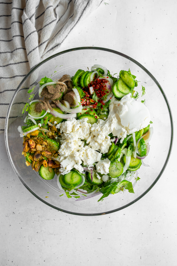 Additional ingredients, including vegan feta, vegan yogurt, and pistachios are added to the mixing bowl.