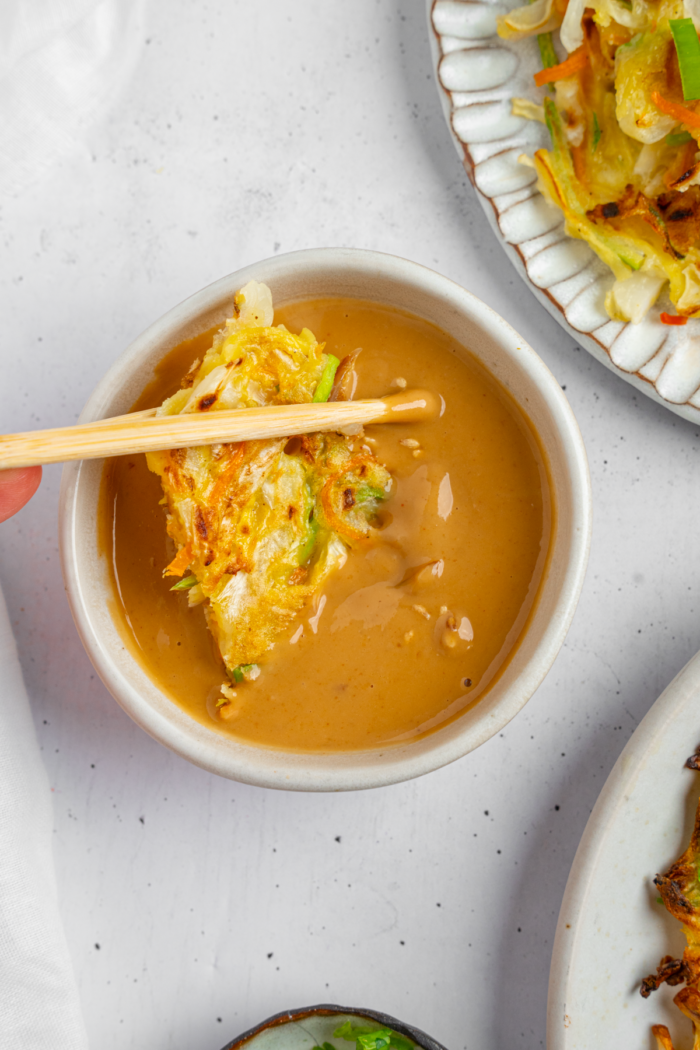 Vegetable pancake being dipped in peanut sauce using chopsticks.