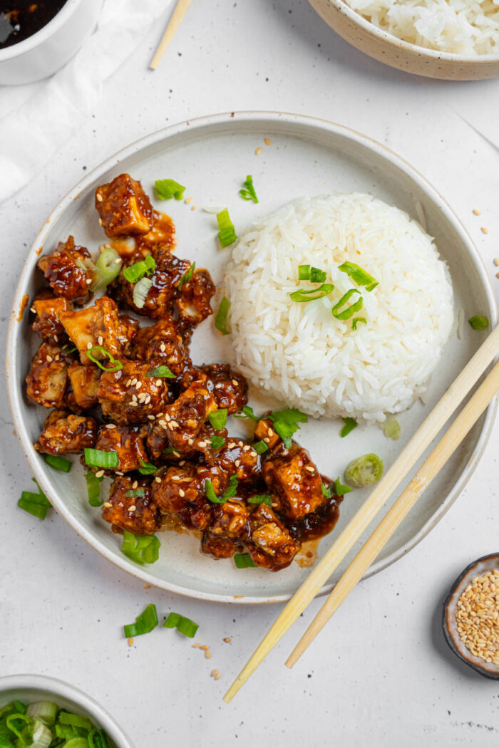 Vegan "honey" garlic tofu served with rice and herbs for garnish, with chopsticks resting on the side of the bowl.