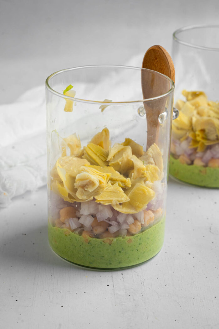 The process of layering the pesto pasta salad in the jar, with the dressing at the bottom, followed by chickpeas and some veggies.