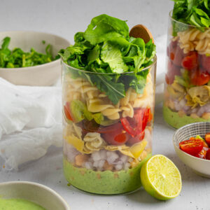 The pesto pasta salad fully assembled in a jar, topped up with fresh greens, with a lime resting against the jar.