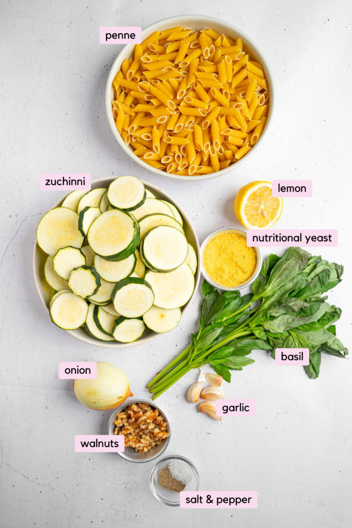 The ingredients for zucchini pesto pasta laid out and labeled on a clear white background.