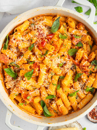 A large ceramic skillet filled with creamy tomato pasta, served with fresh basil and vegan cheese shot from above.